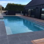 Luxurious in-ground swimming pool with modern black house exterior, grey tiled surround, and wooden deck, perfect for outdoor living and relaxation in Bermuda.