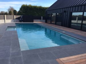 Luxurious in-ground swimming pool with modern black house exterior, grey tiled surround, and wooden deck, perfect for outdoor living and relaxation in Bermuda.