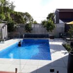 Bright blue in-ground pool with glass fencing, surrounded by a modern outdoor patio with lounge chairs, umbrella, and lush greenery, perfect for Bermuda outdoor living and pool renovations.