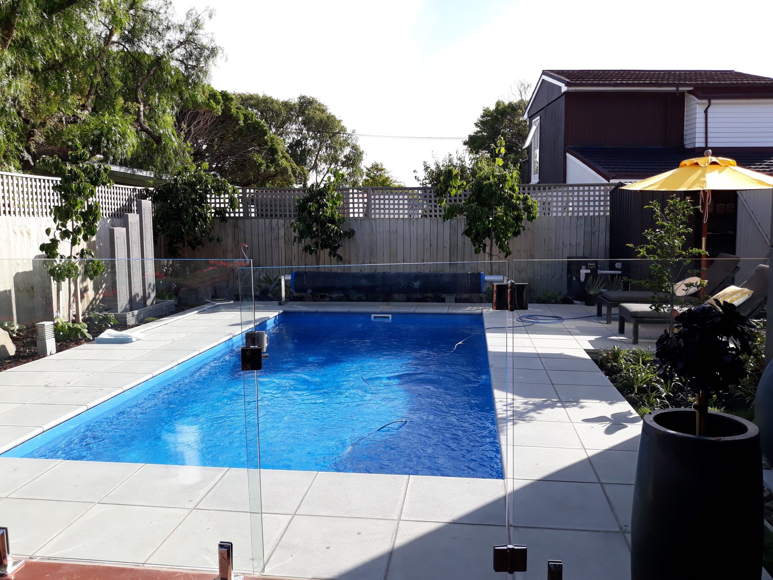 Bright blue in-ground pool with glass fencing, surrounded by a modern outdoor patio with lounge chairs, umbrella, and lush greenery, perfect for Bermuda outdoor living and pool renovations.