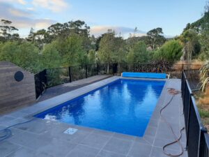Modern private swimming pool with sleek blue water, surrounded by a stylish grey tiled deck and black safety fencing, nestled within lush green tropical landscape in Bermuda.