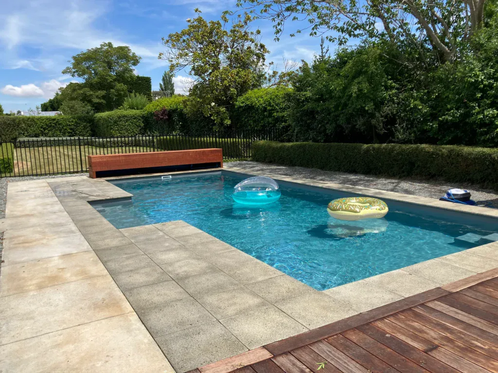 Vibrant blue swimming pool with inflatable floaties, surrounded by a mix of stone and wooden decking, garden hedge, and lush greenery in Bermuda. Perfect for relaxing summer days.