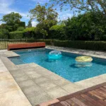 Vibrant blue swimming pool with inflatable floaties, surrounded by a mix of stone and wooden decking, garden hedge, and lush greenery in Bermuda. Perfect for relaxing summer days.