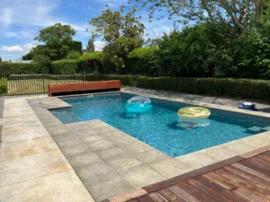 Vibrant blue swimming pool with inflatable floaties, surrounded by a mix of stone and wooden decking, garden hedge, and lush greenery in Bermuda. Perfect for relaxing summer days.