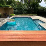 Bright backyard swimming pool with lounging floats, surrounded by modern stone tiles and lush greenery, perfect for relaxing and entertaining. Bermudan Pools offers custom pool designs and outdoor living solutions.