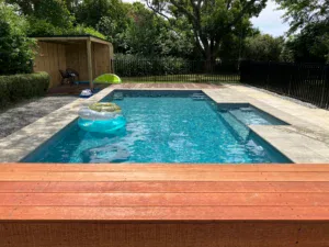Bright backyard swimming pool with lounging floats, surrounded by modern stone tiles and lush greenery, perfect for relaxing and entertaining. Bermudan Pools offers custom pool designs and outdoor living solutions.
