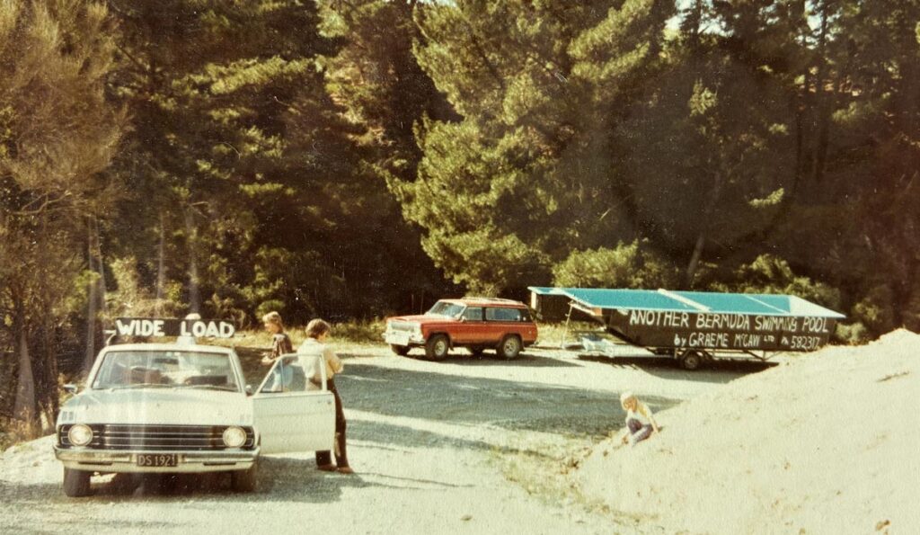 Wide load signage on a vintage vehicle parked near Bermuda Pools, with children and trees in the background, showcasing early Bermuda pool construction and family leisure scenes.