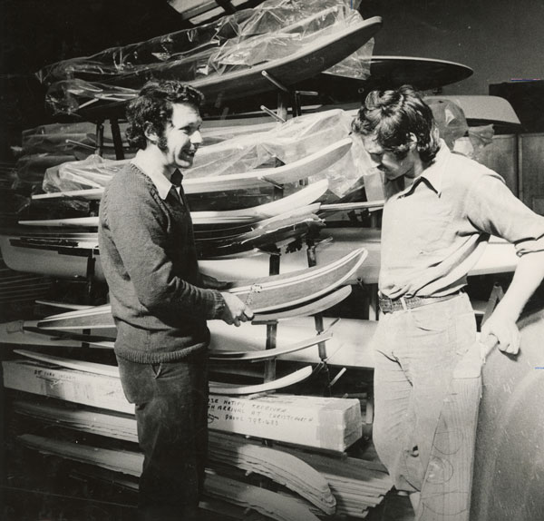 Vintage photo of two men inspecting surfboards in a warehouse, embodying the history of Bermuda surfing culture. Ideal for showcasing surfboard manufacturing or surf sport heritage.